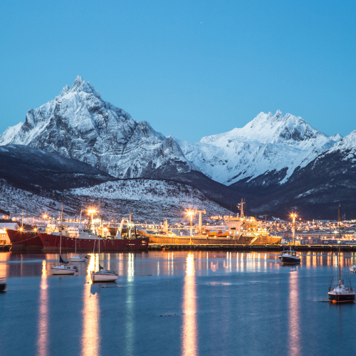 Pacote de viagem para Ushuaia exibindo o porto iluminado ao anoitecer com barcos ancorados no Canal Beagle e as imponentes montanhas nevadas da Patagônia Argentina ao fundo, destacando o turismo de imersão e ecoturismo cultural em uma expedição internacional exclusiva rumo ao Fim do Mundo.