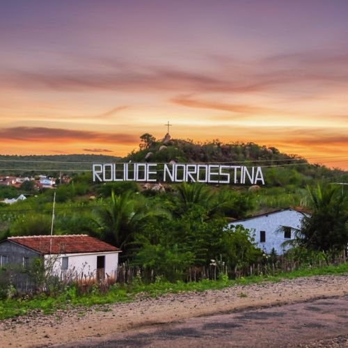 Letreiro branco com a frase Roliúde Nordestina no topo de uma colina verde sob céu de fim de tarde na estrada de Cabaceiras, Paraíba.