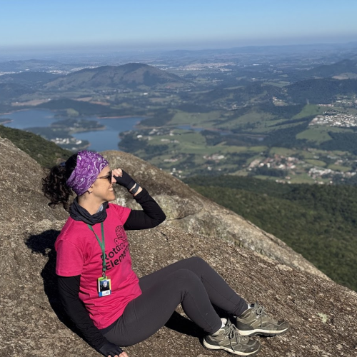 Mulher sentada no topo de uma montanha contemplando um vale verde em trilha de ecoturismo.